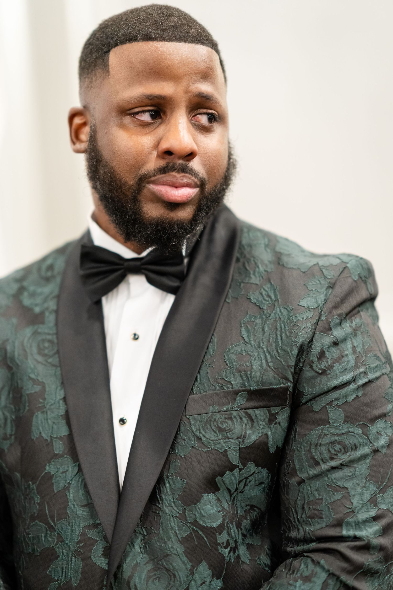 Groom poses in sophisticated emerald and black floral jacquard tuxedo with bow tie.