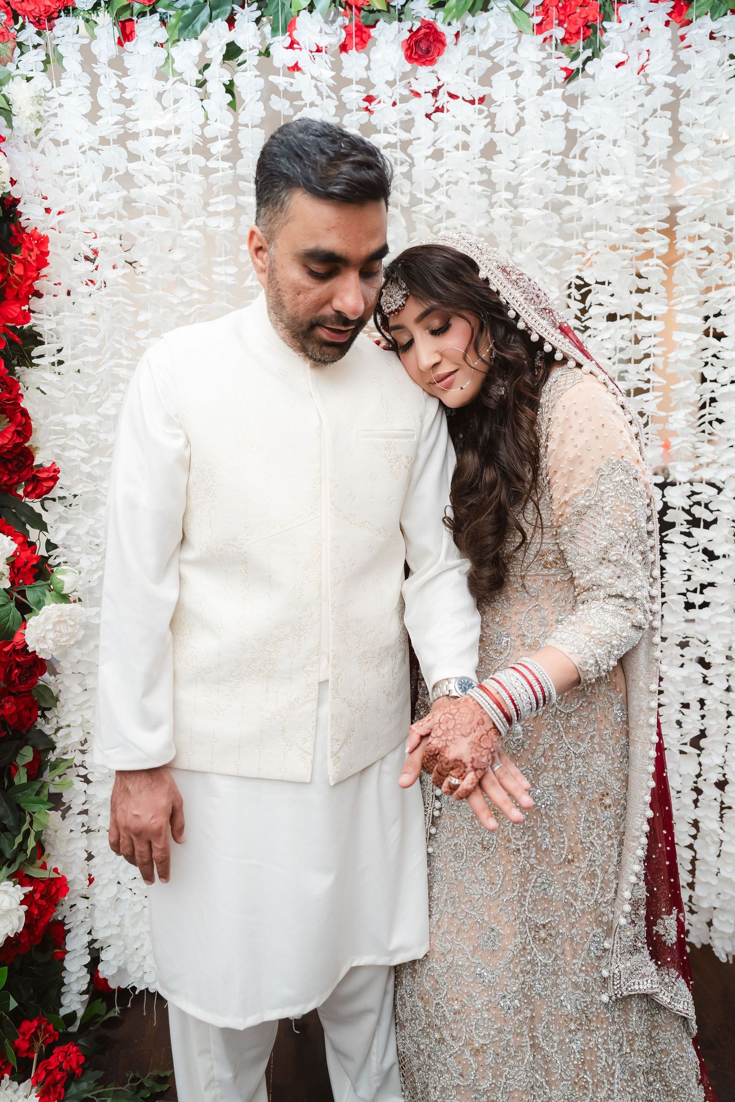 A couple in white and silver wedding attire poses against a floral backdrop with hanging decorations.