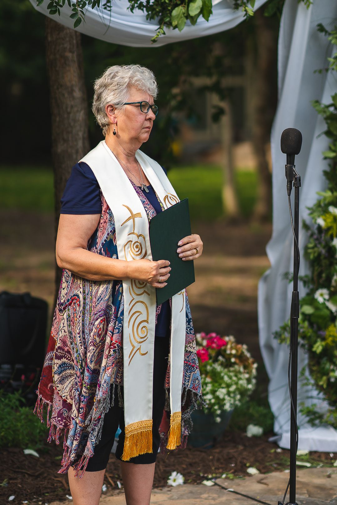 An officiant in a navy dress and ceremonial stole stands at an outdoor wedding ceremony.