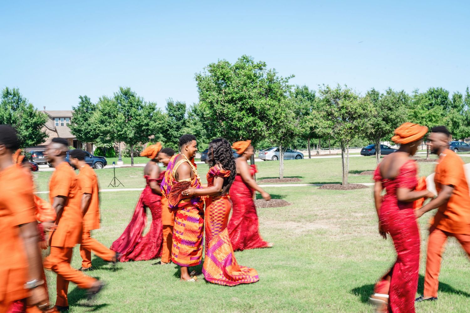 Large wedding party in red and orange attire posing together outdoors on a sunny day.