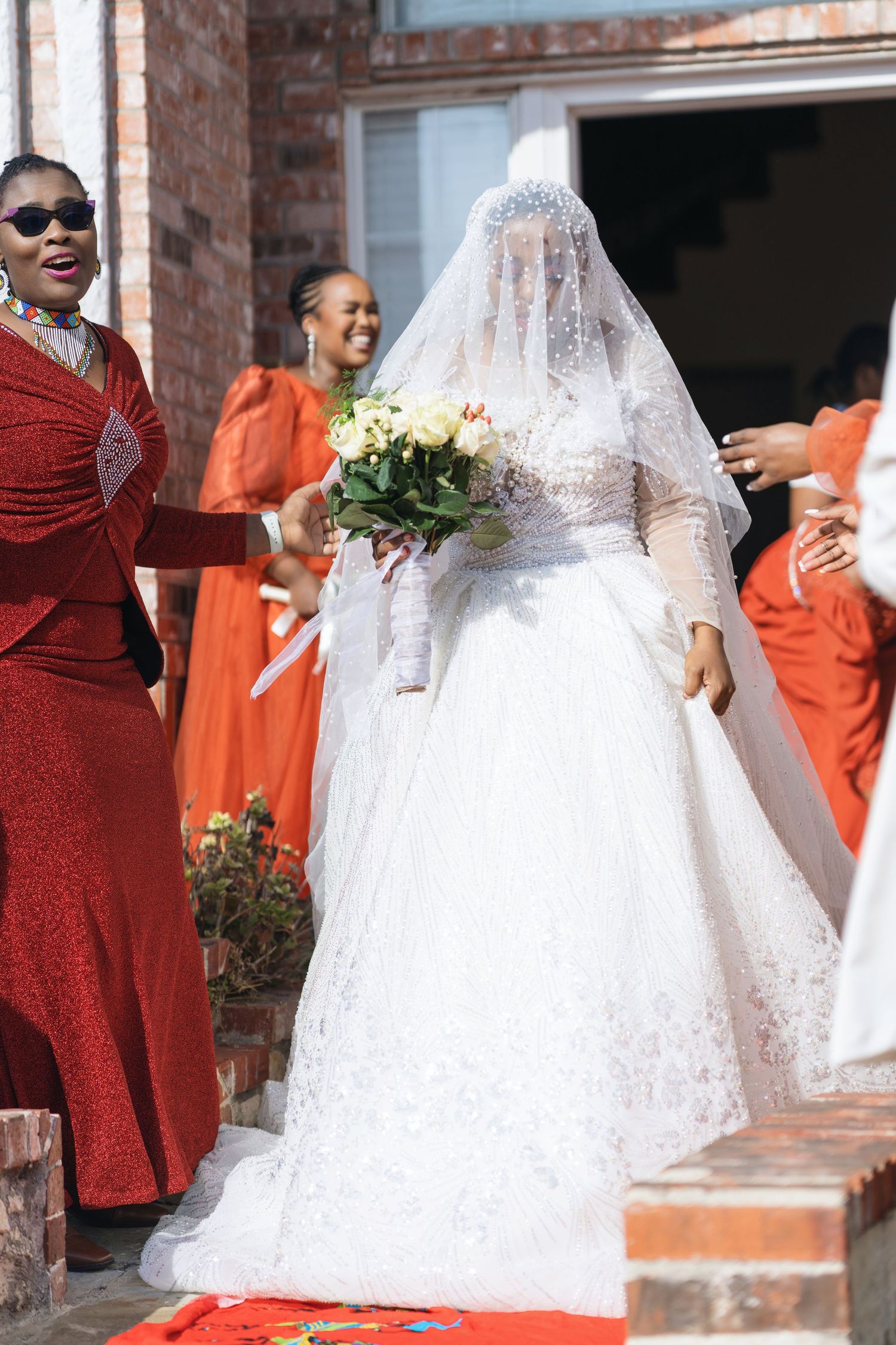 Wedding party members in vibrant orange dresses stand with the bride in her white lace gown outdoors.