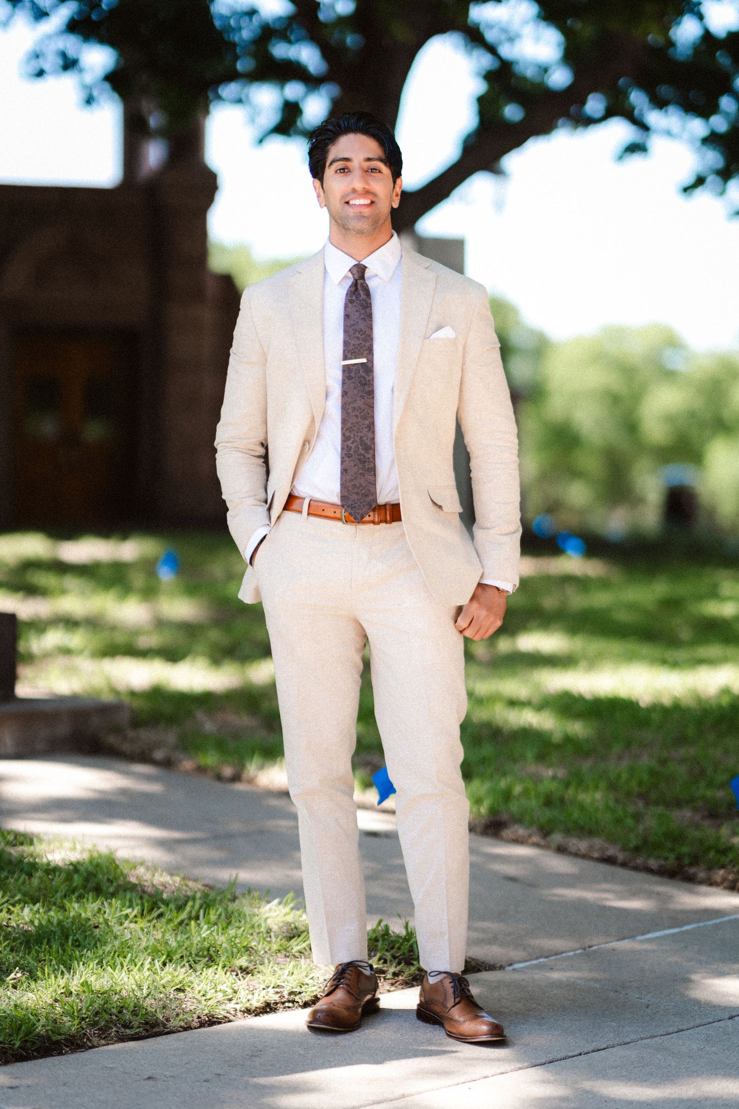 A person in a light beige suit and brown tie stands on a sunlit outdoor walkway.