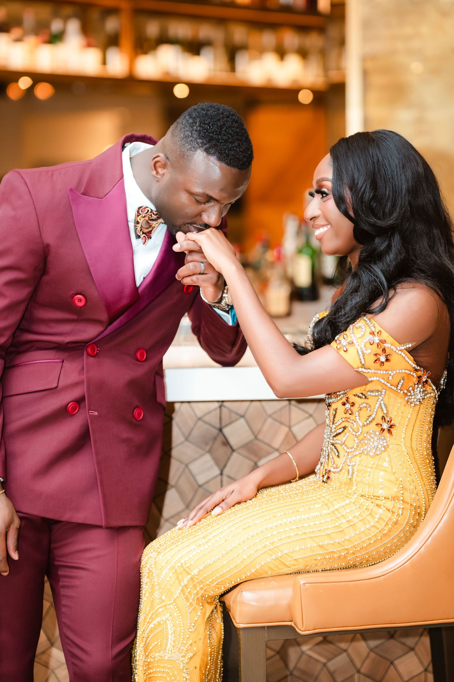 A couple poses together, with one wearing a burgundy suit and the other in a golden gown.