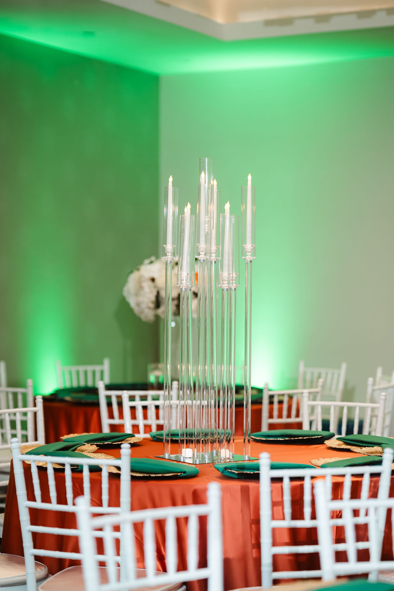 Wedding reception venue with white chiavari chairs and orange tablecloths illuminated by elegant green uplighting.