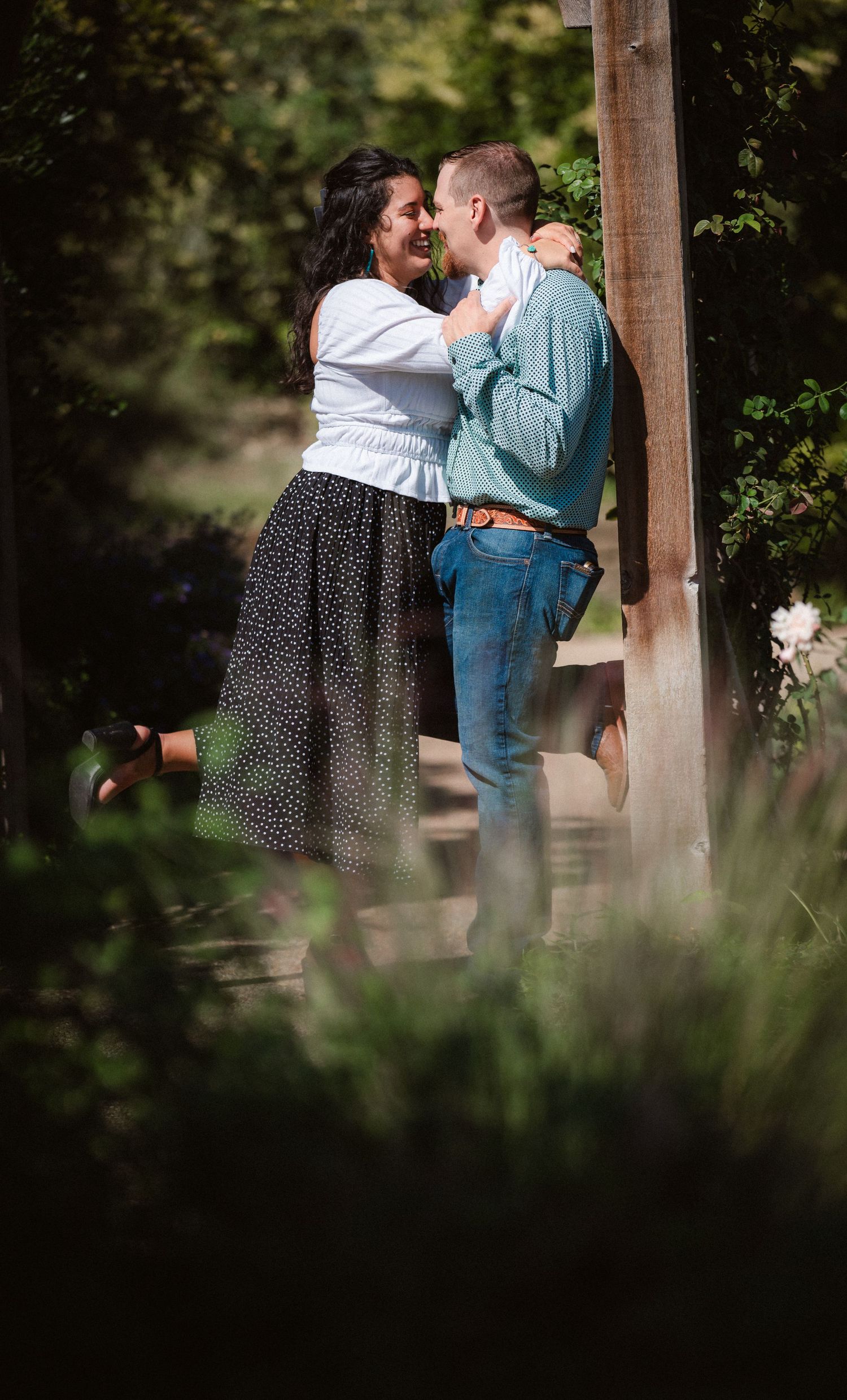 A romantic couple embraces under a wooden gazebo in a lush garden setting.