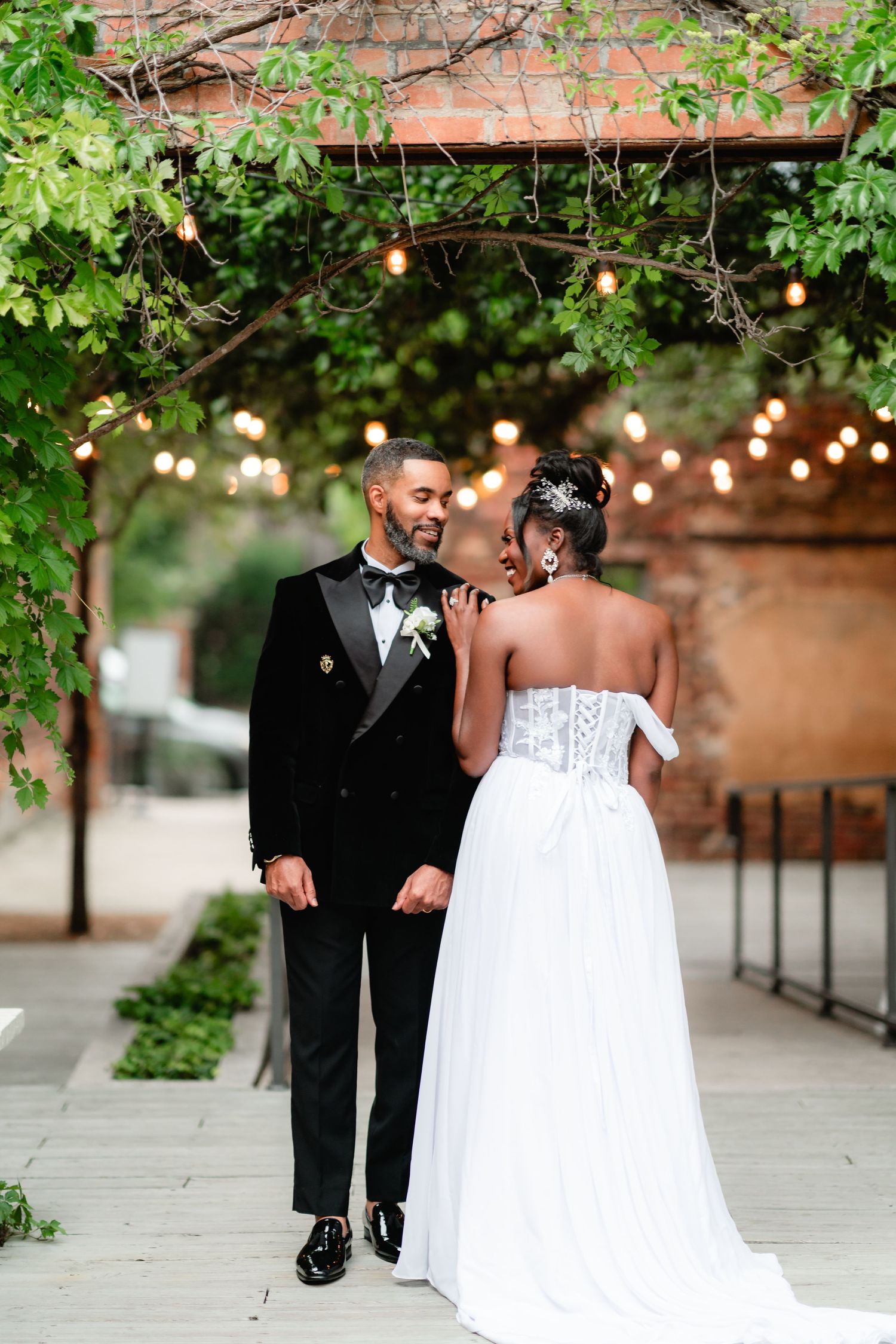 A wedding couple shares an intimate moment outdoors under string lights and greenery.
