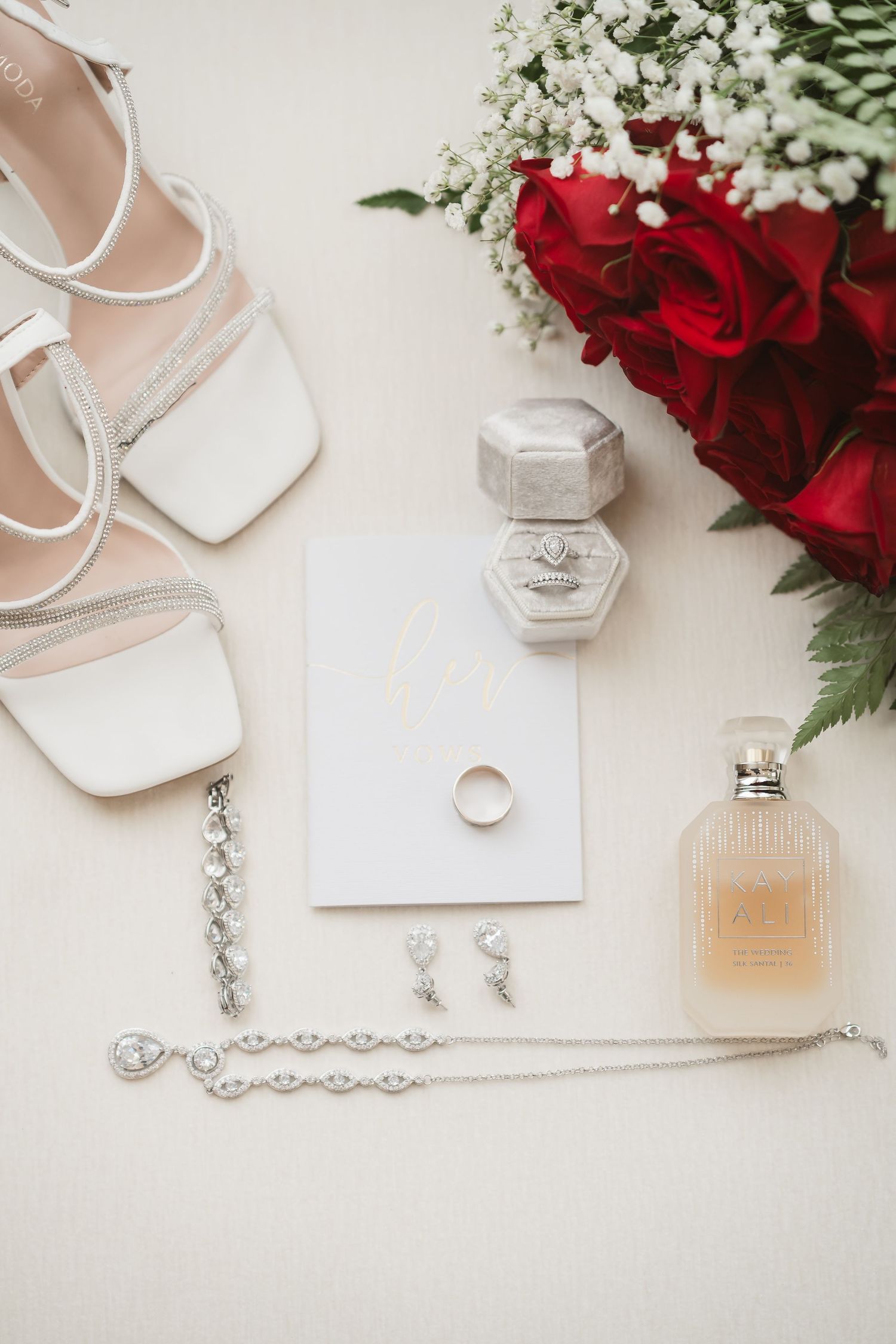 Wedding accessories including white shoes, jewelry, perfume, and red roses arranged on a light background.