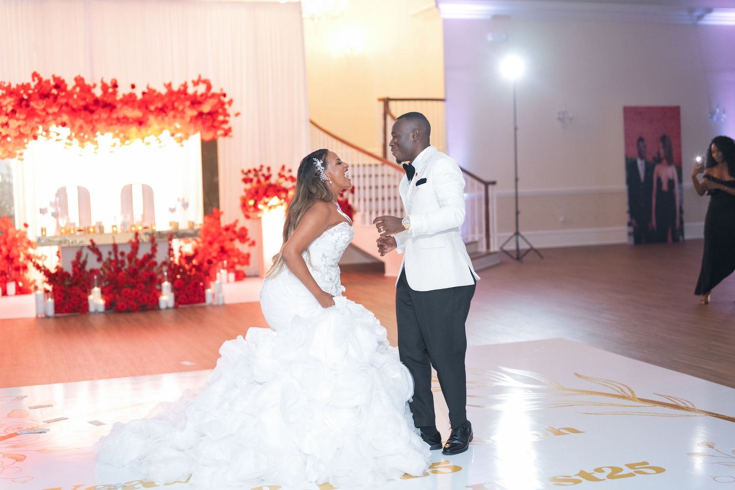Wedding couple dancing in an elegant white reception space with red floral decorations.