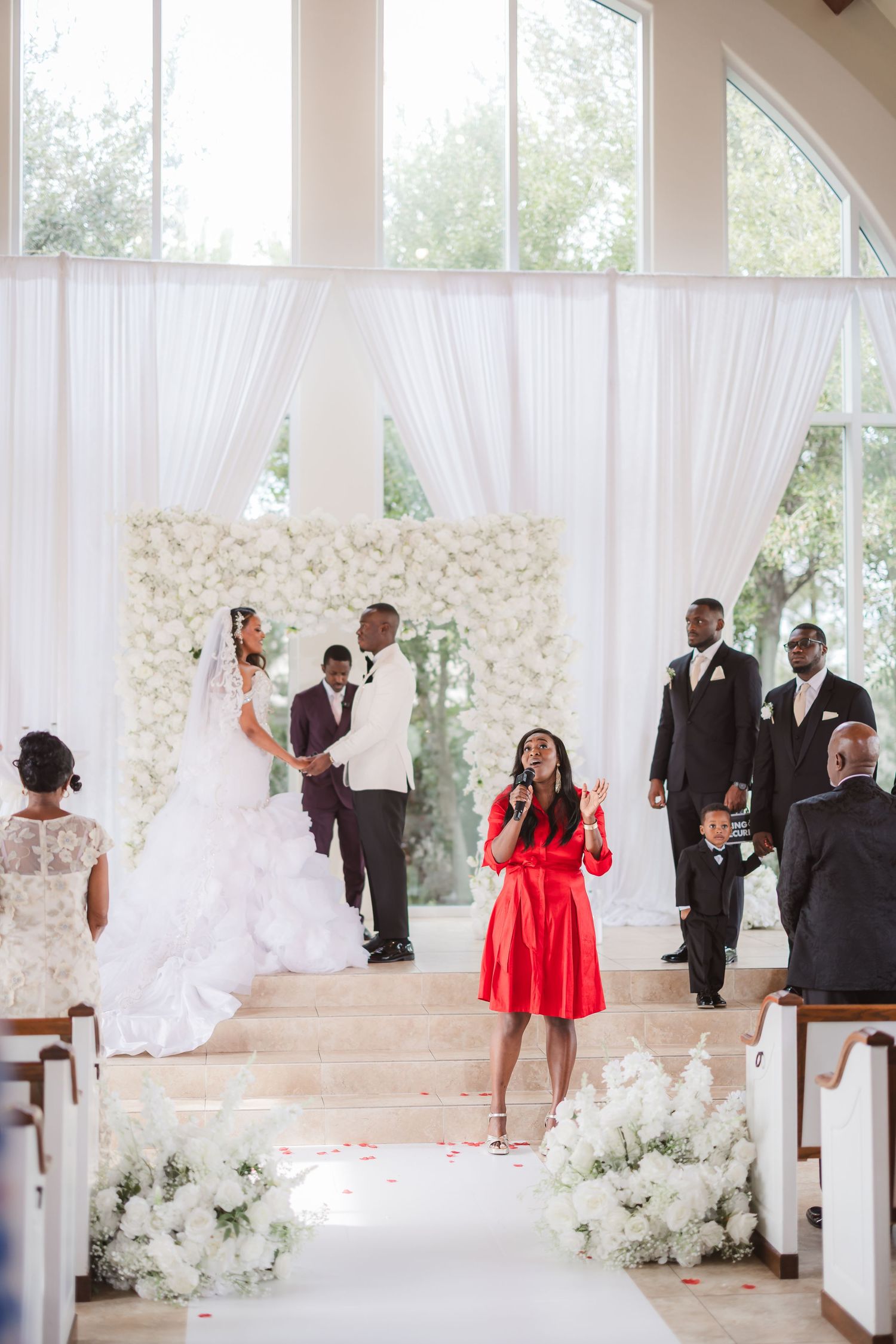 Wedding ceremony venue with white floral decorations, pews, and sheer curtains in an elegant white room.