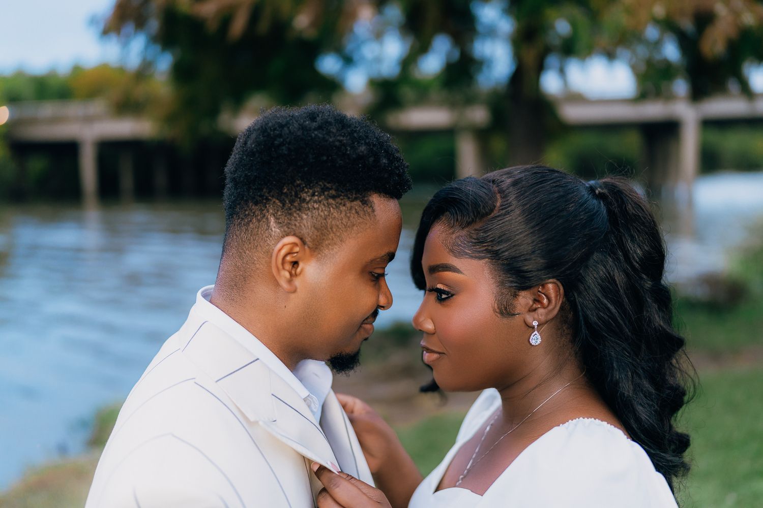 A romantic couple in white outfits embraces outdoors near a waterfront during sunset.