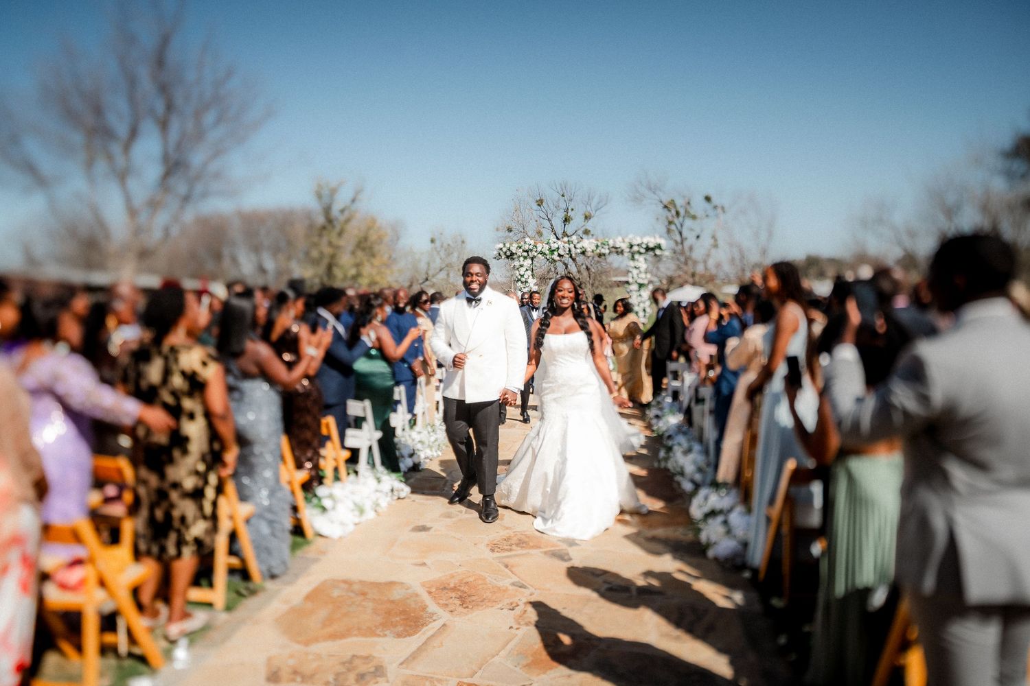 A crowd of wedding guests lines an outdoor ceremony aisle during a sunny afternoon celebration.
