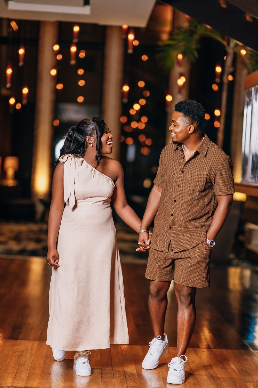 A couple in coordinating beige and brown outfits dances on a wooden floor under decorative lighting.