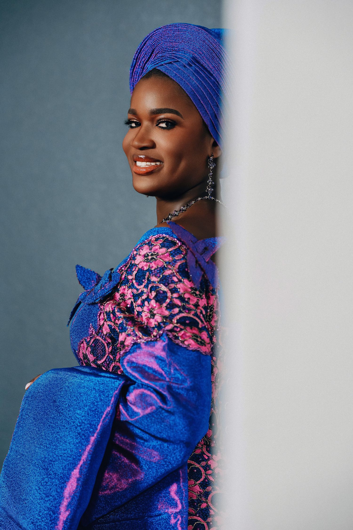 Woman in vibrant blue and pink traditional African dress poses elegantly against grey wall.