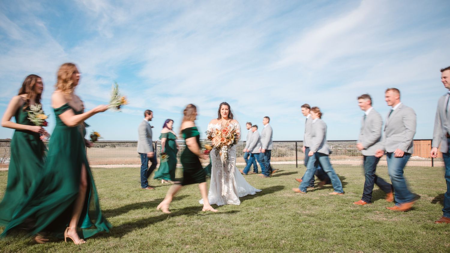 Wedding guests celebrate on a grassy field under blue skies during an outdoor reception.