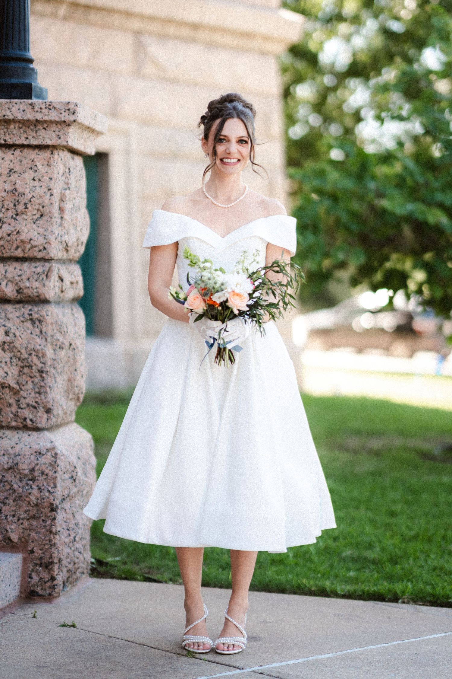 A tea-length white wedding dress and bouquet are showcased against a stone building backdrop.