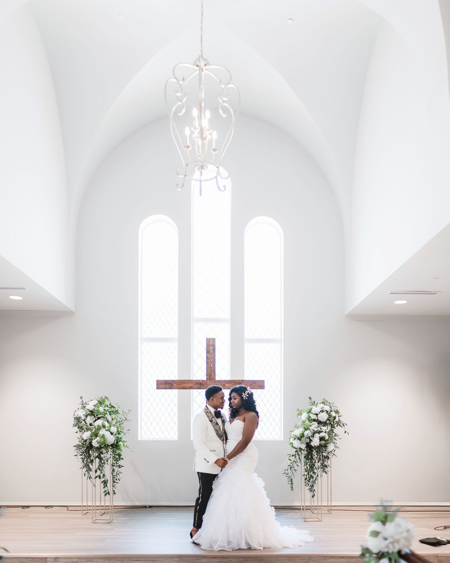 A romantic wedding ceremony scene in an elegant white chapel with arched windows and floral decorations.