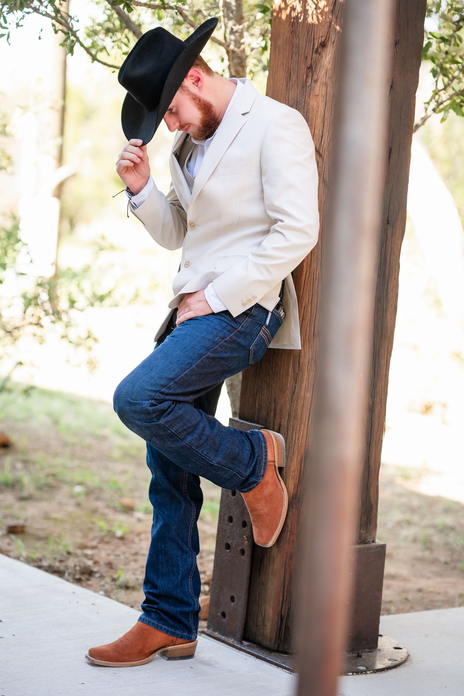 A man in a white blazer and cowboy hat leans against a wooden post.