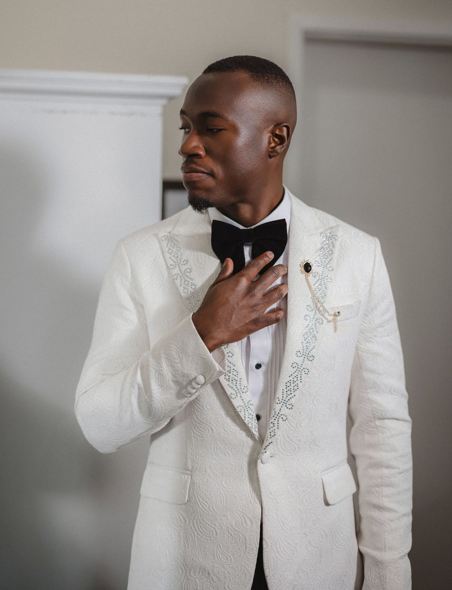 Groom in an elegant white embroidered tuxedo jacket poses thoughtfully before the wedding ceremony.