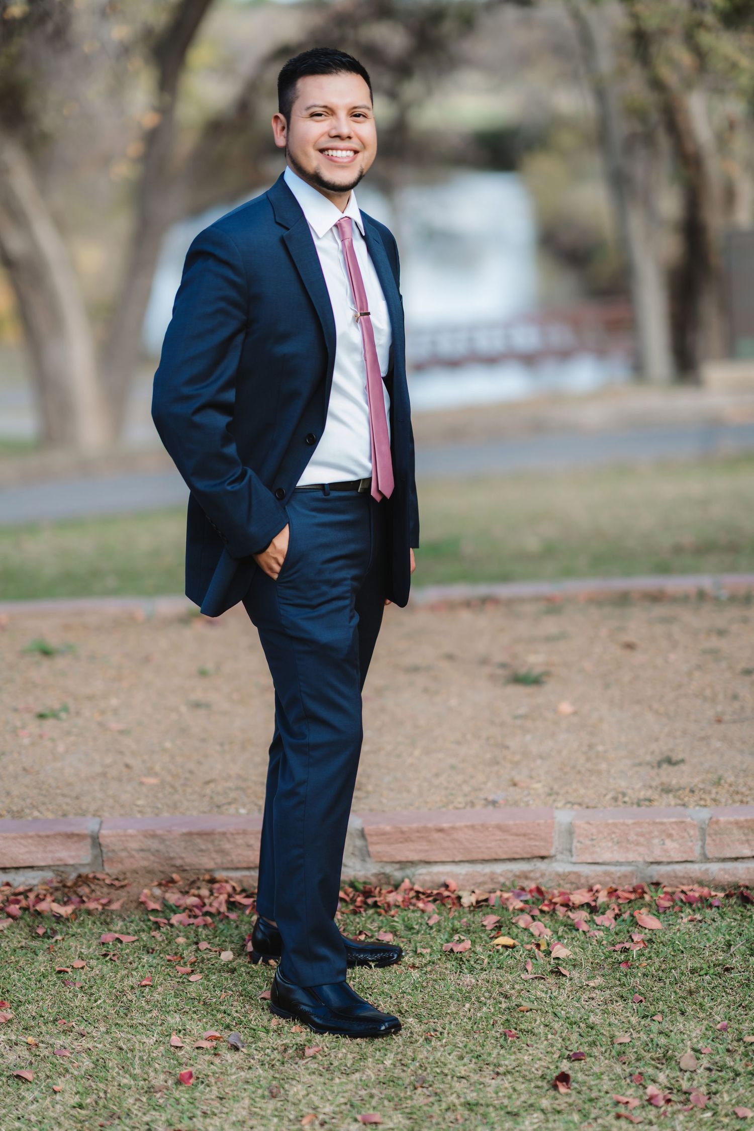 A person in a navy suit and pink tie poses playfully in a series of outdoor portraits on grass and fallen leaves.