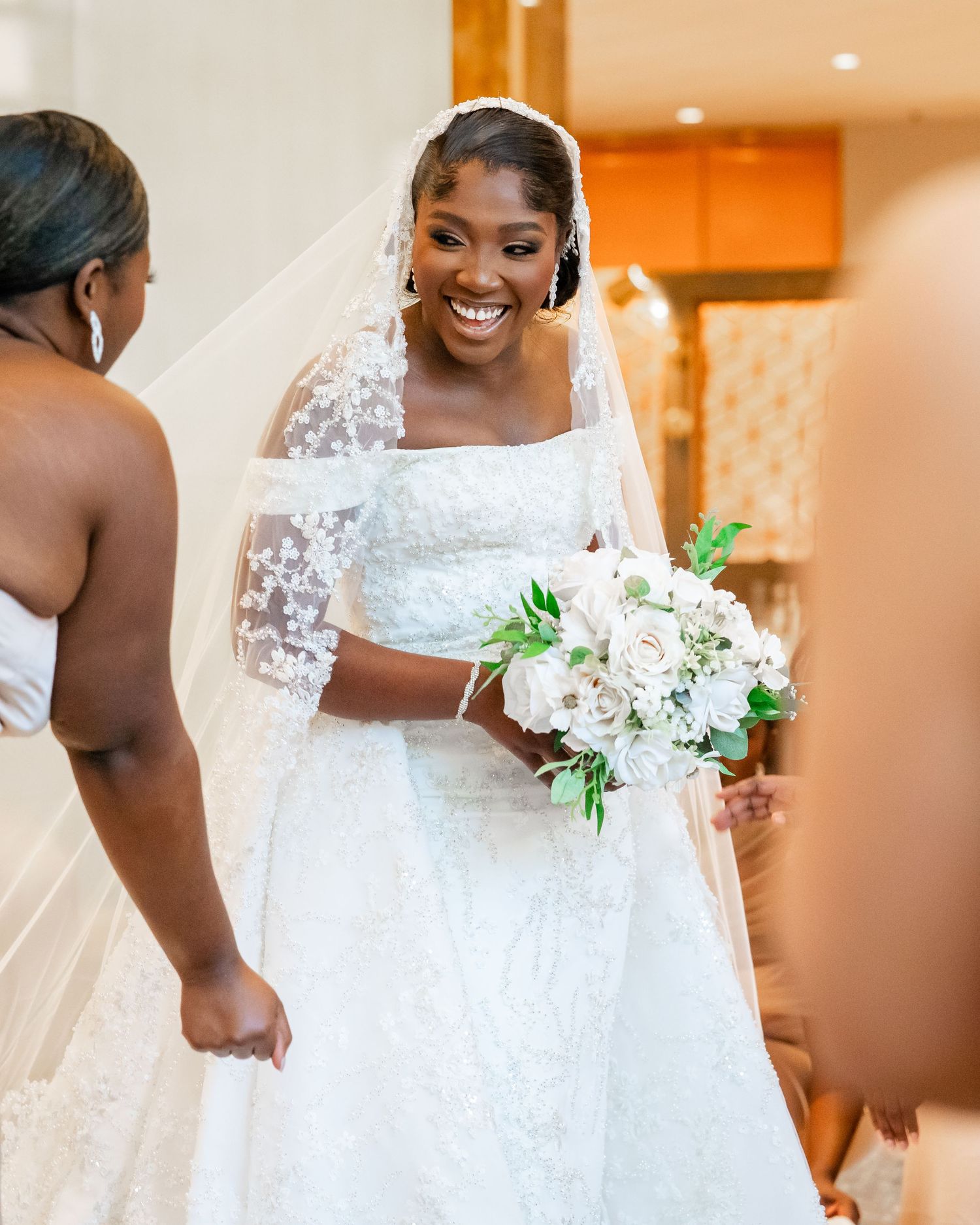 A bride in a white lace wedding gown holds a bouquet of white flowers during preparation.