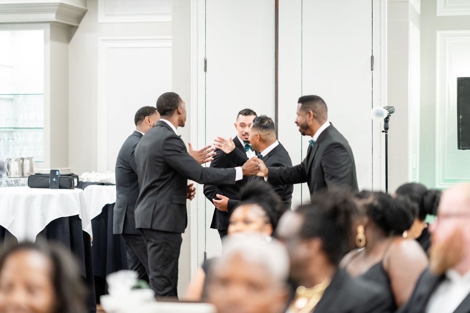 A group of wedding guests in formal attire gather in a bright indoor venue.