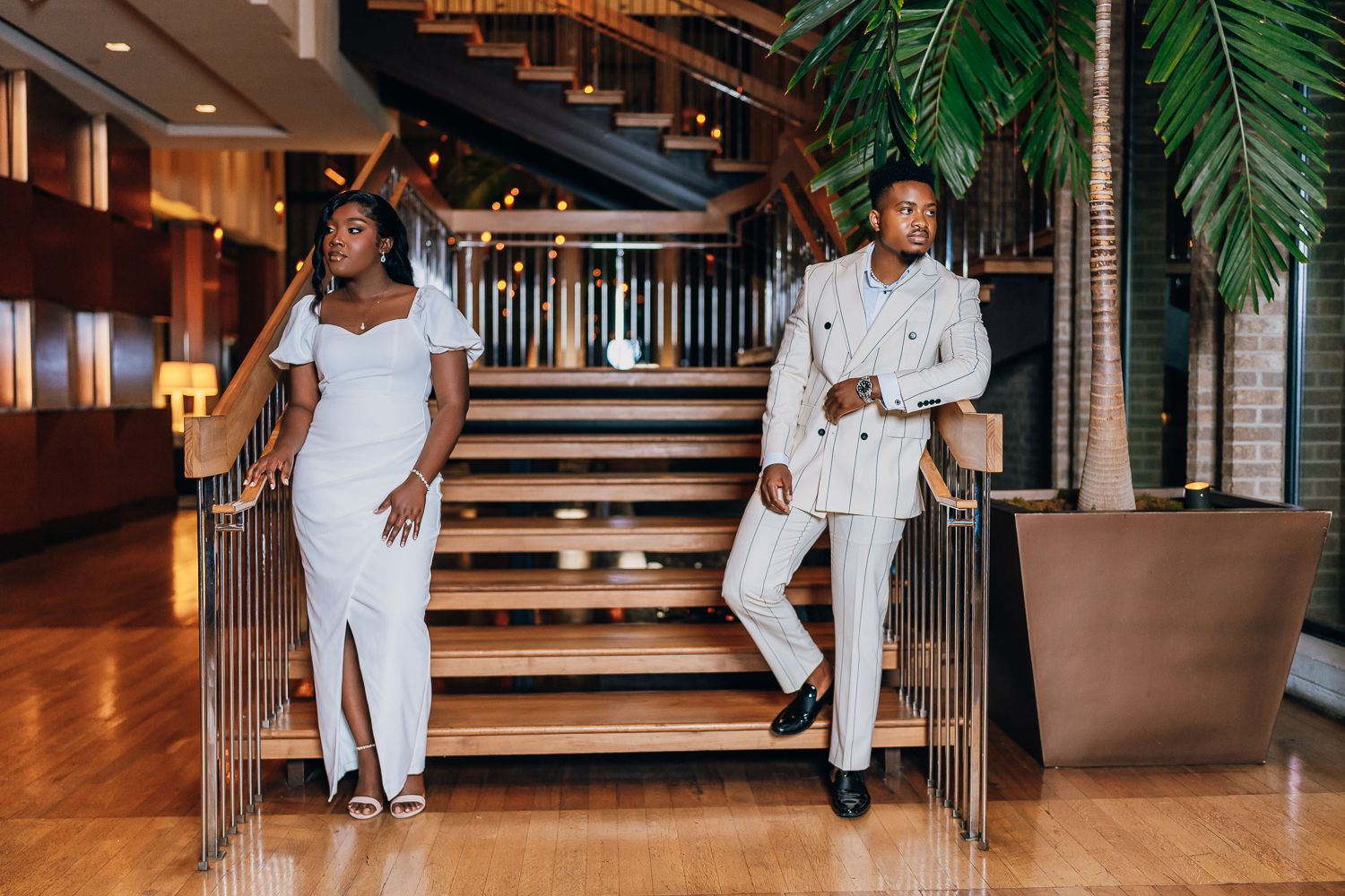 A couple in matching all-white outfits dances playfully in an elegant indoor venue with palm decor.