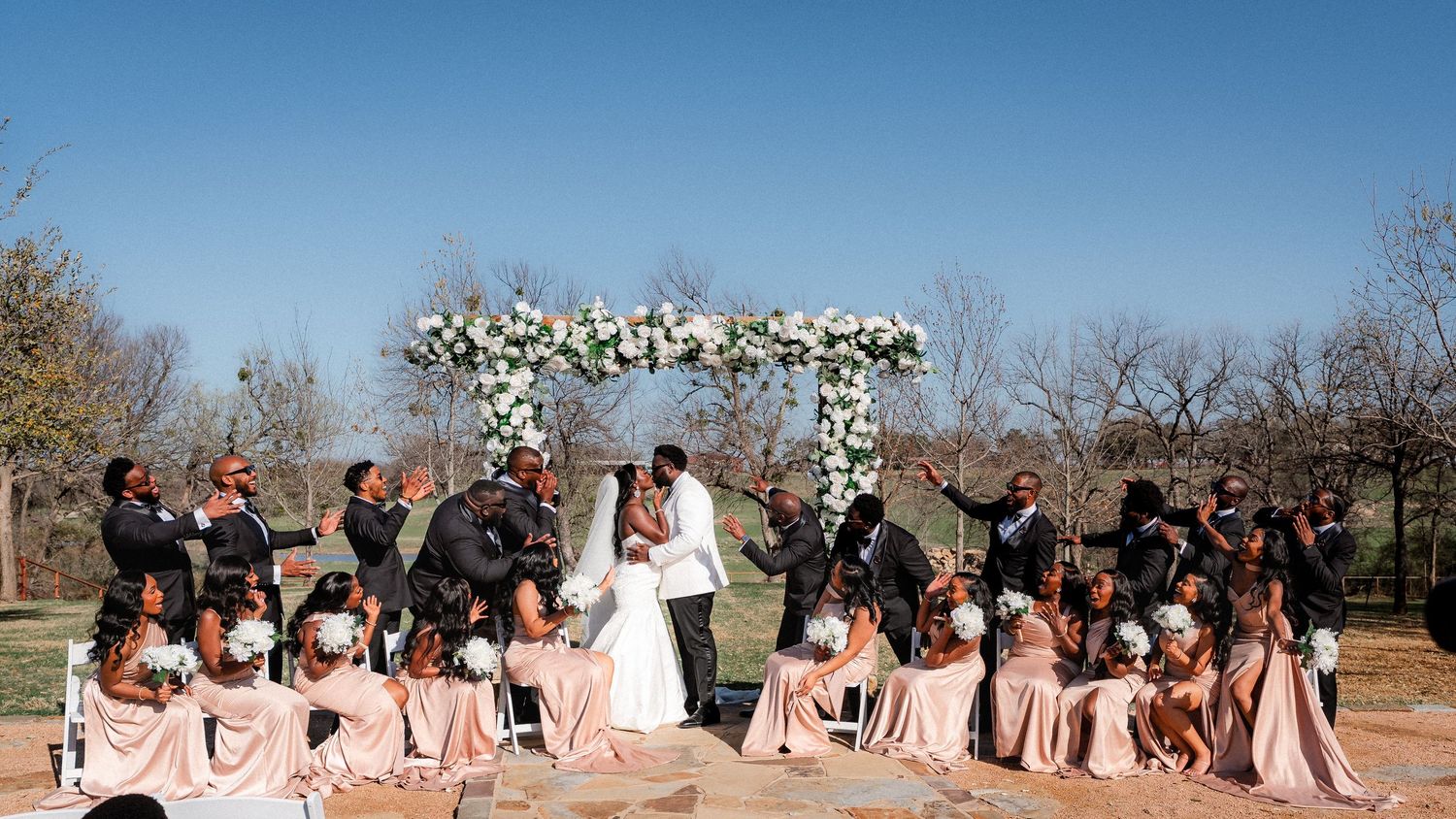 Wedding party poses outdoors on a sunny day with floral arch decorations and pink bridesmaids dresses.