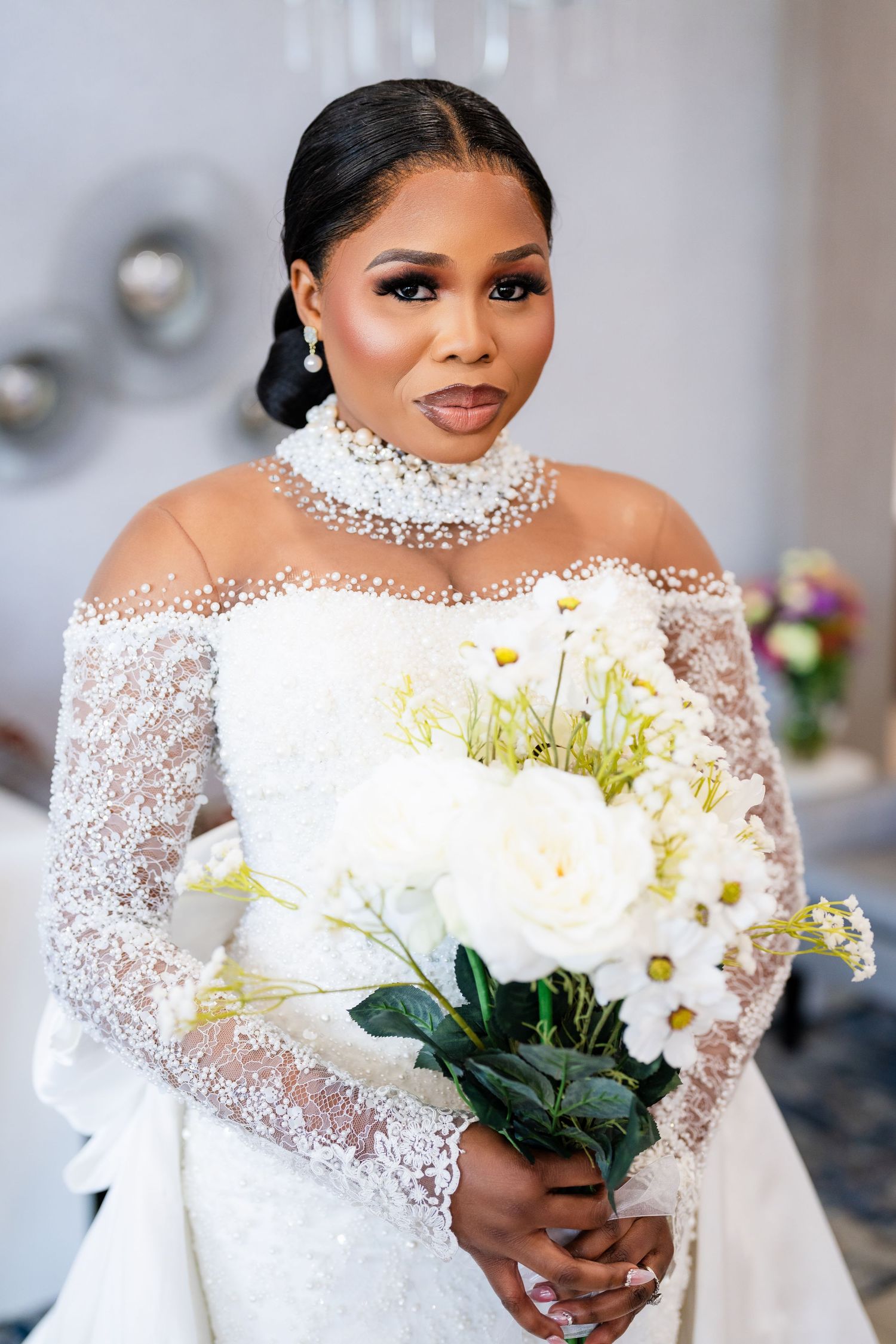 A bride in an off-shoulder lace gown holds a white floral bouquet against a light background.