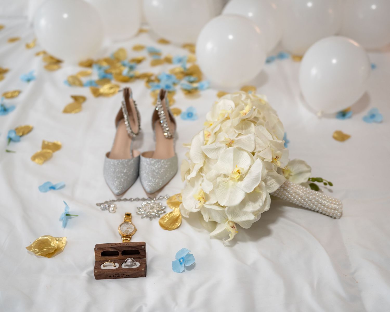 Wedding rings rest on white fabric with scattered gold and blue confetti and delicate white flowers.