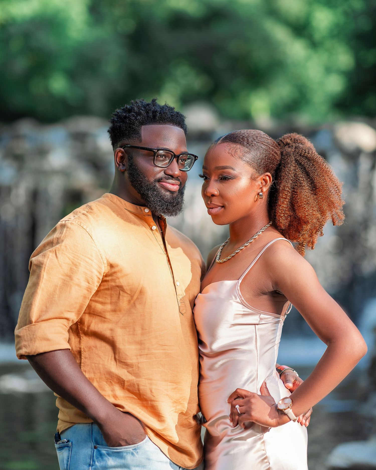 A romantic couple poses together outdoors, with one wearing an orange shirt and the other in a light colored top.