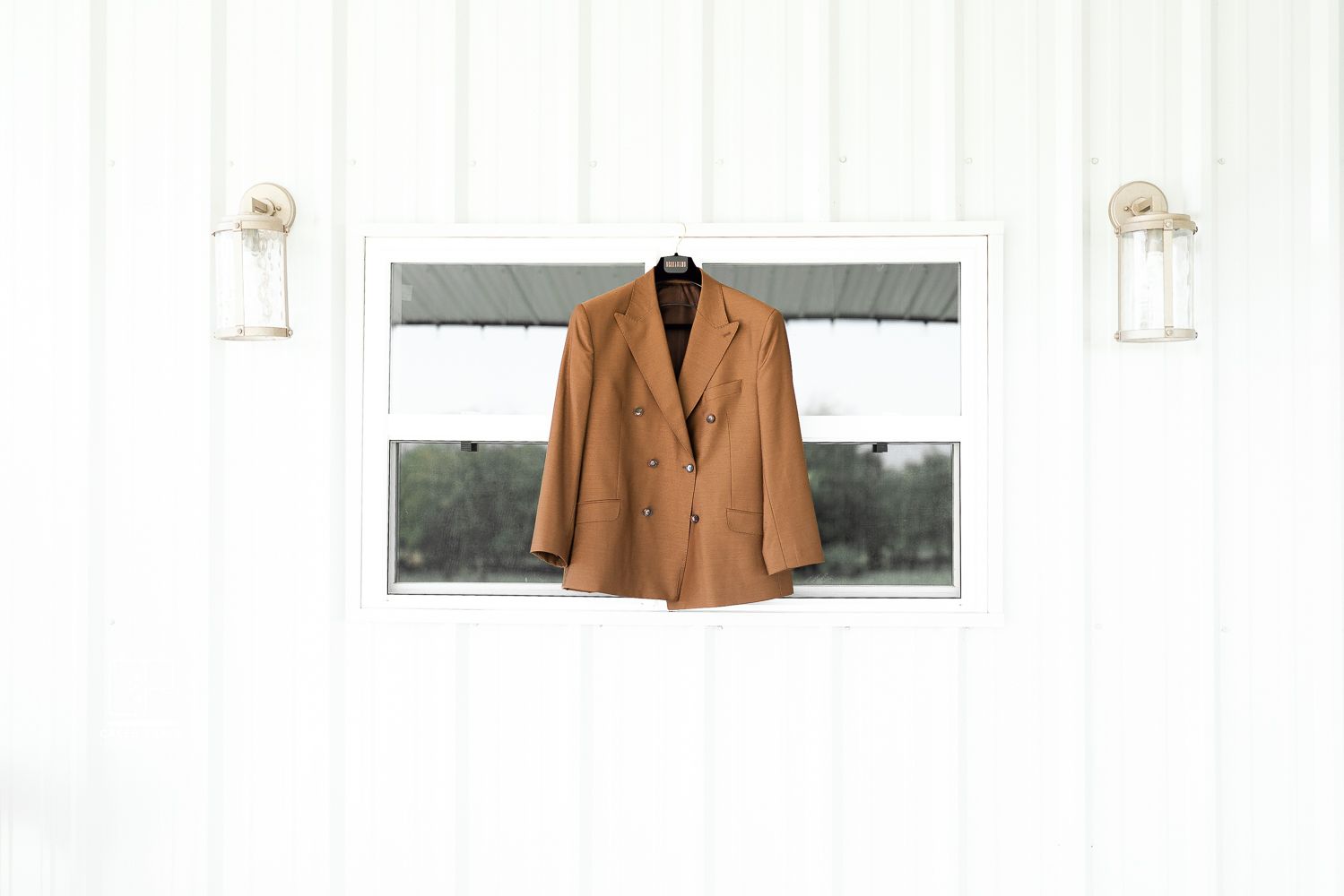 Tan suit jacket hanging on white wall between two wall sconces for wedding day preparation.