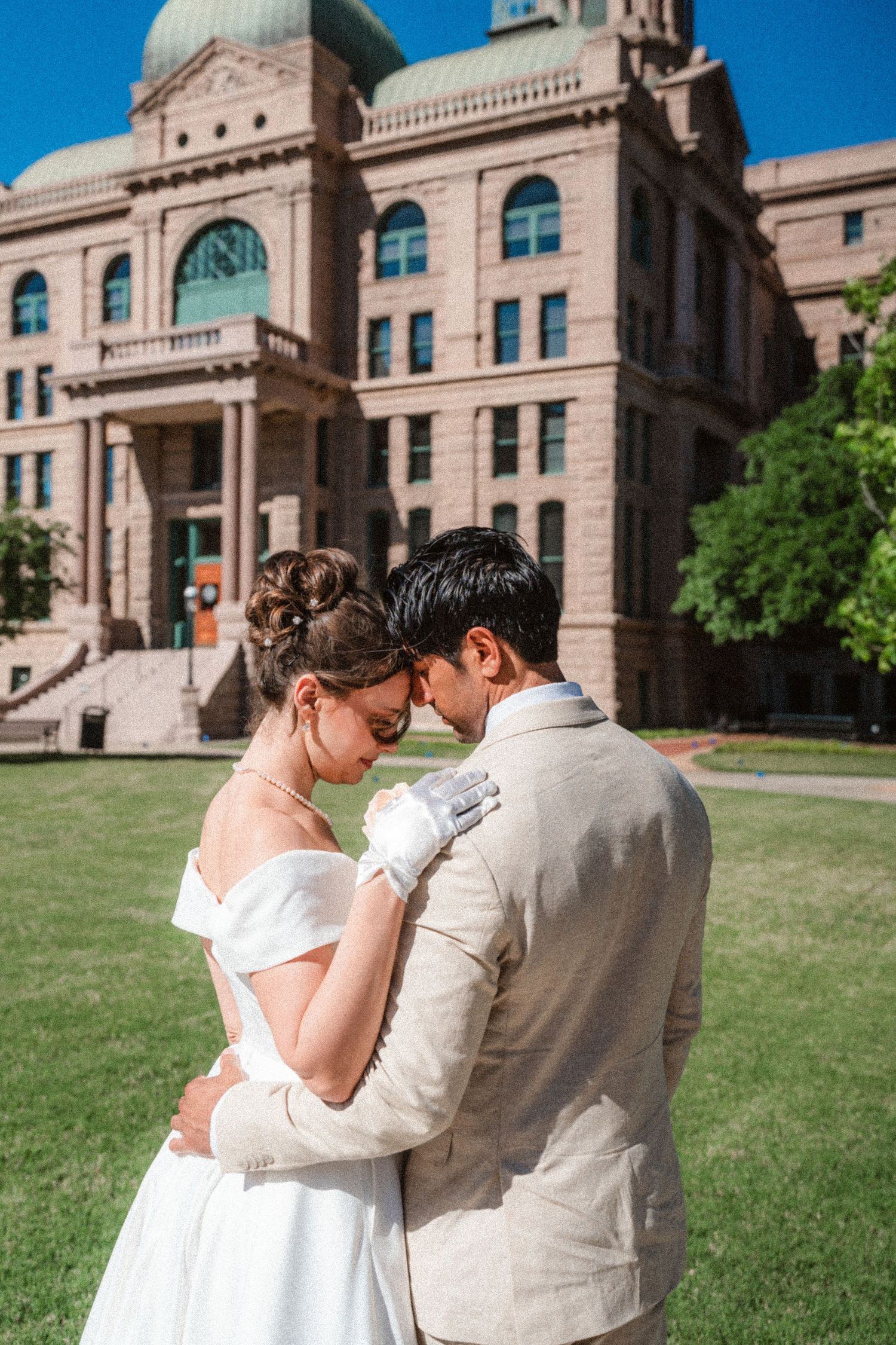 A wedding couple embraces in front of a grand historic building with beige stone architecture.