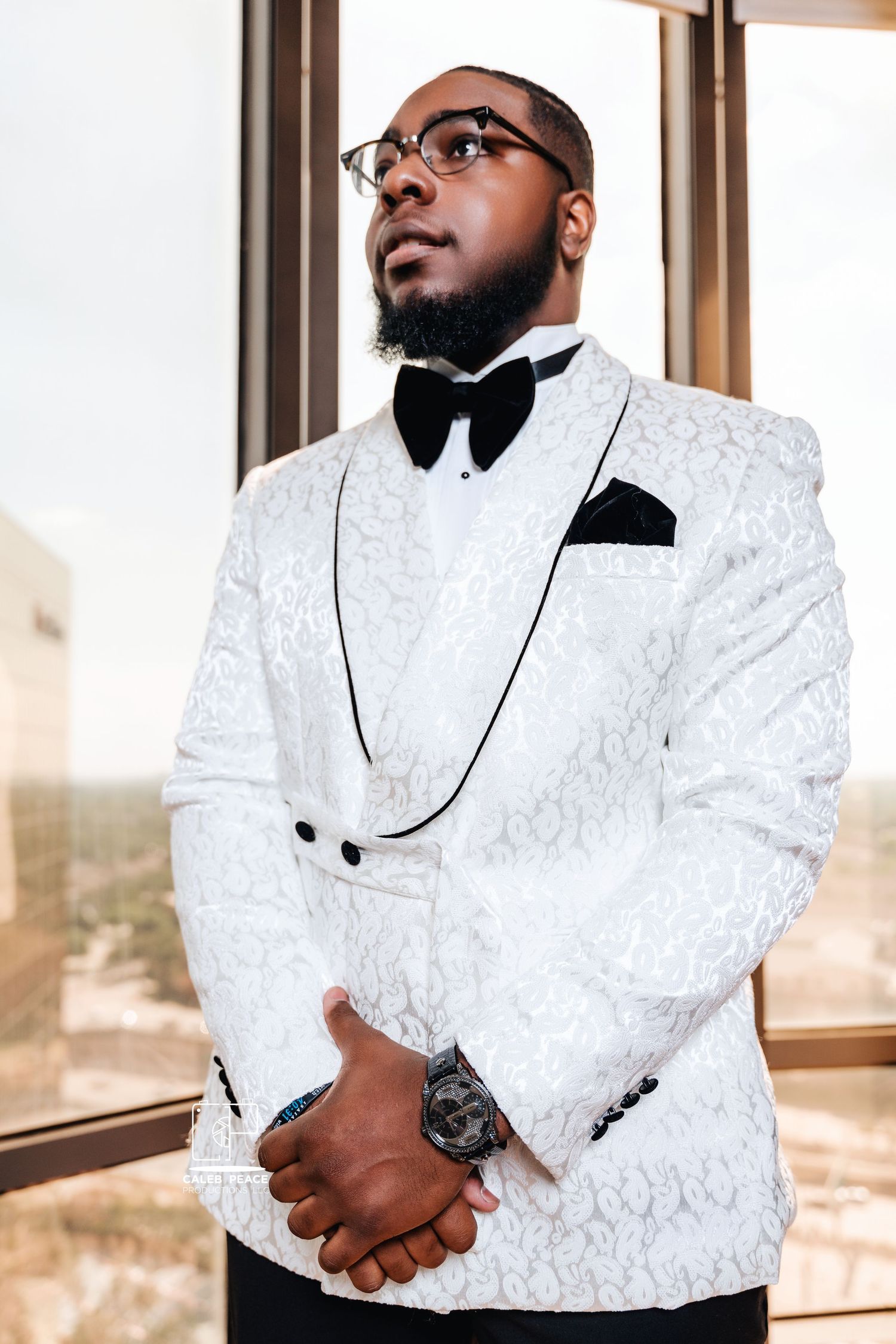 Stylish groom wearing a white textured tuxedo jacket with black bow tie and glasses in formal wedding attire.