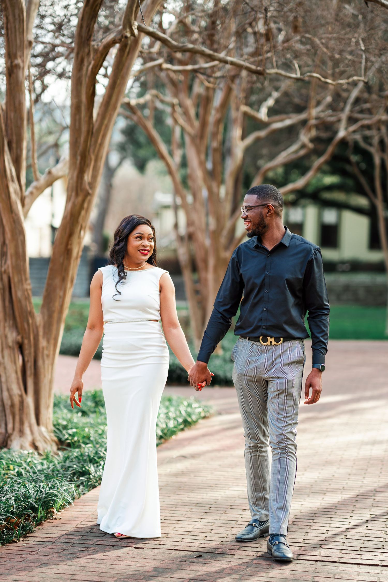 A couple walks together on a brick path surrounded by crepe myrtle trees in a garden setting.