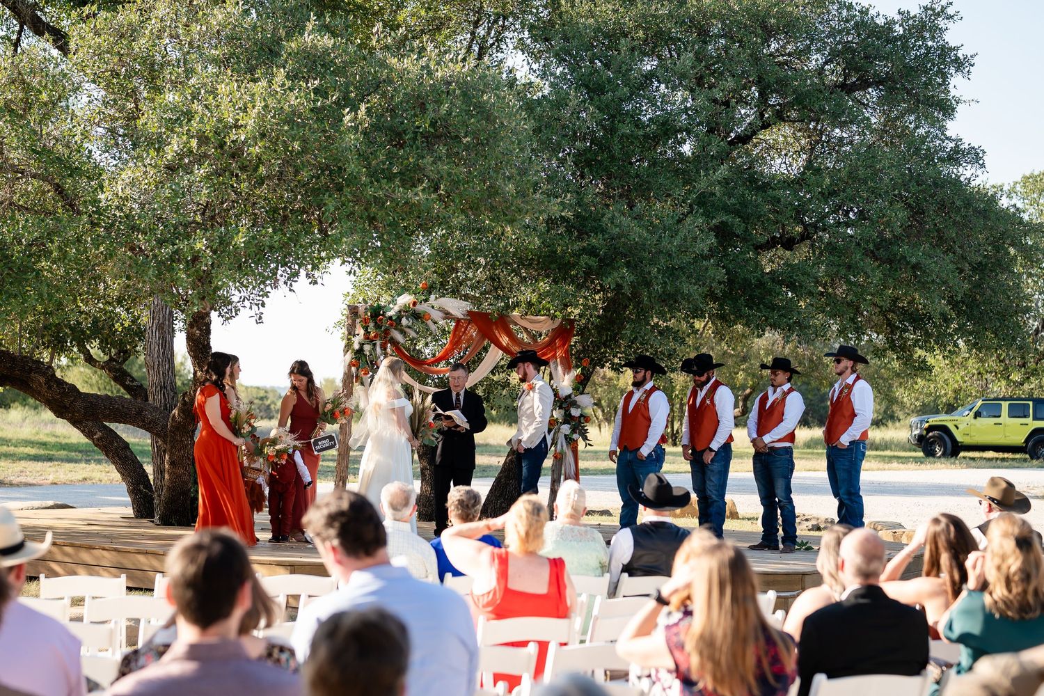 Wedding ceremony in progress with bridal party in red dresses and guests seated outdoors.