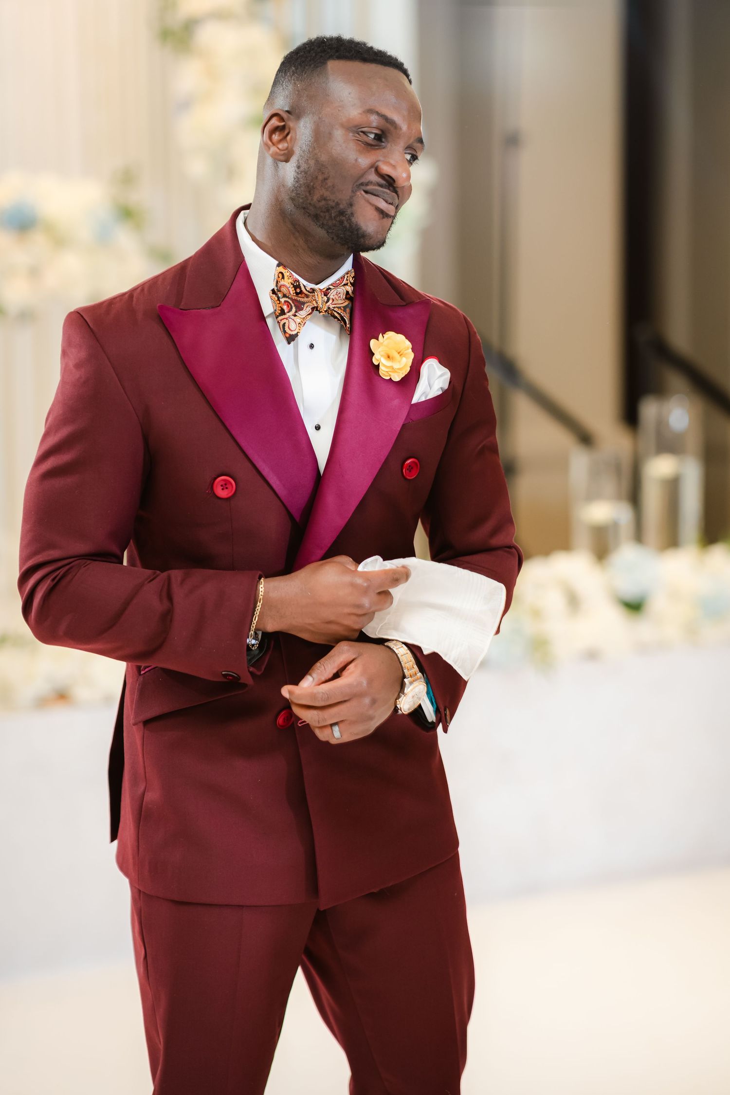 Groom in burgundy tuxedo with white boutonniere at elegant wedding venue.