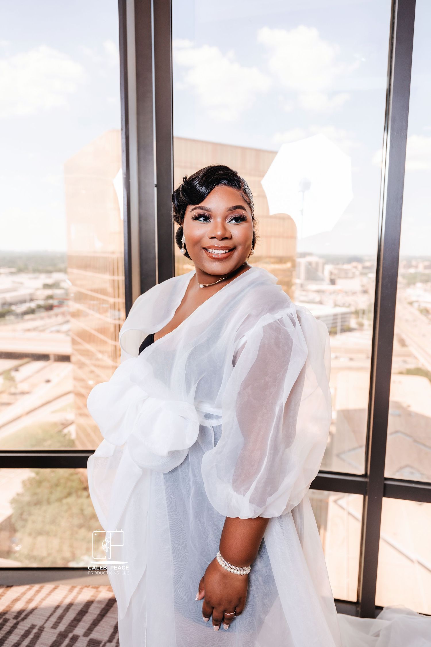 A bride in a white wedding dress with dramatic puff sleeves poses elegantly by large windows with a scenic view.