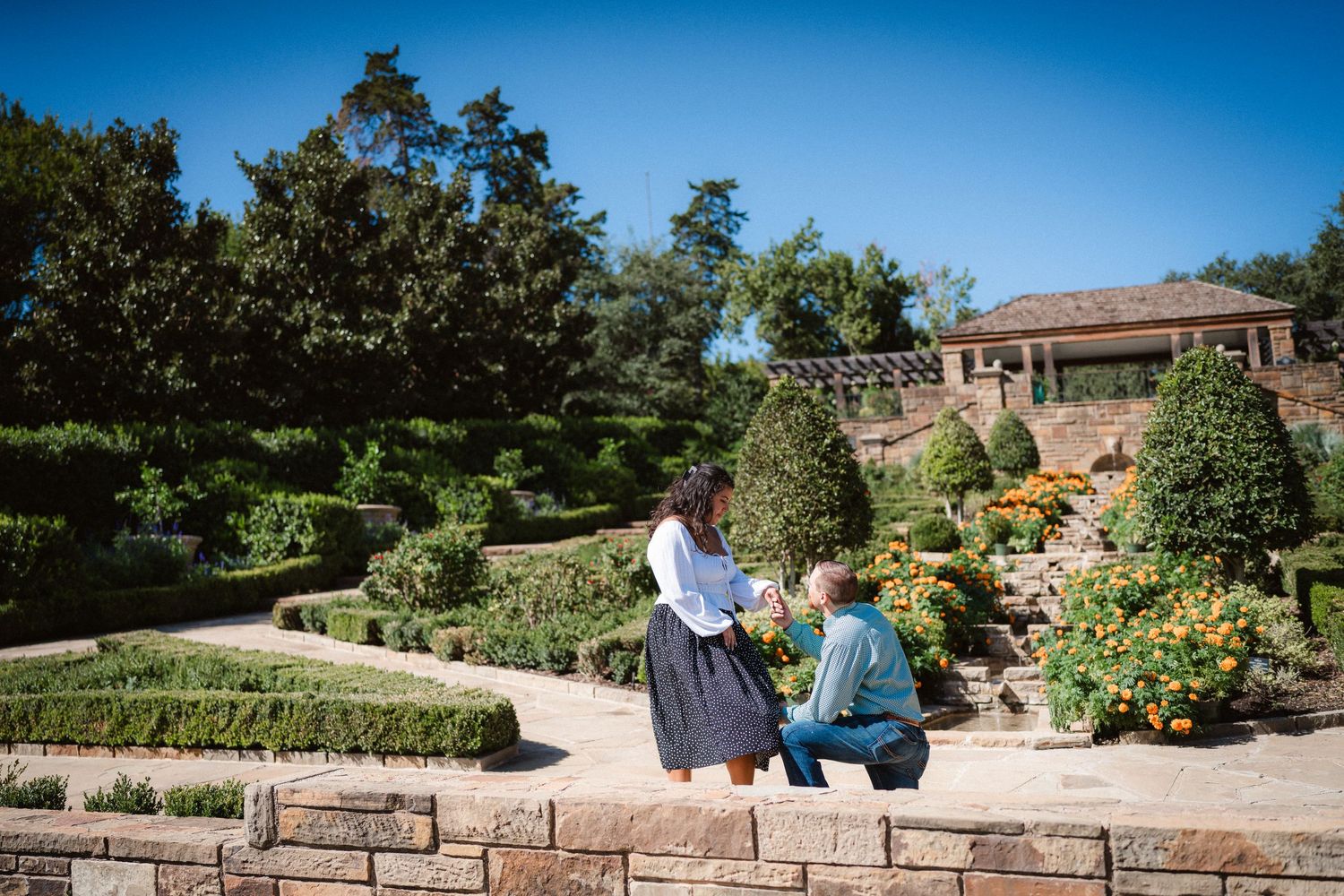 Panoramic view of a well-maintained garden with stone walls, benches, and blooming flowers on a sunny day.