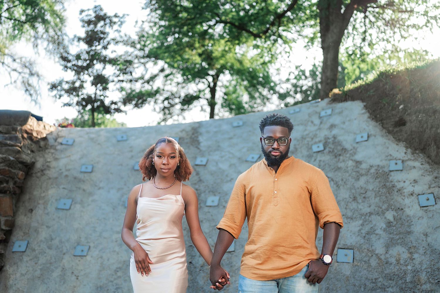 A couple holds hands while posing against a stone wall in an outdoor setting.