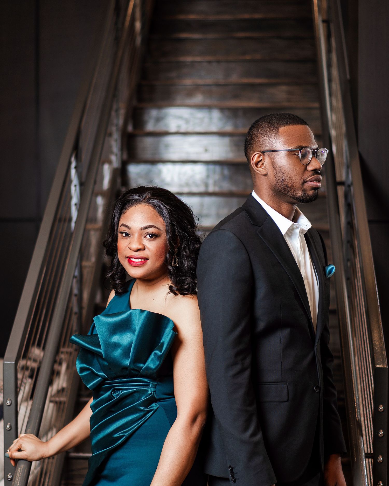 Couple in formal attire poses against dark wooden staircase backdrop with dramatic lighting.