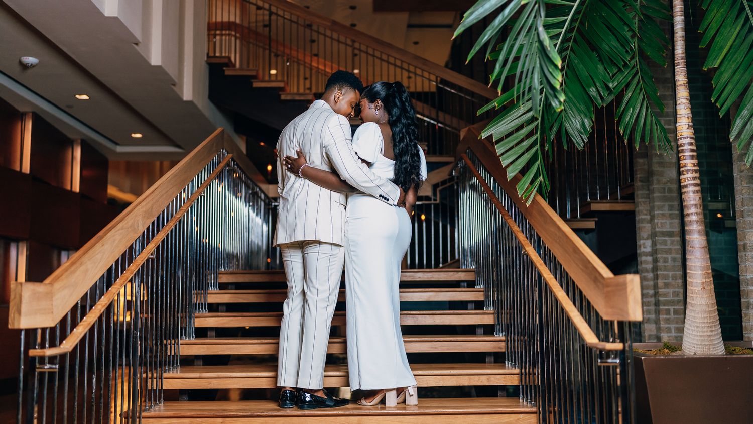 A couple poses together on wooden stairs with palm trees and ambient lighting creating a romantic atmosphere.