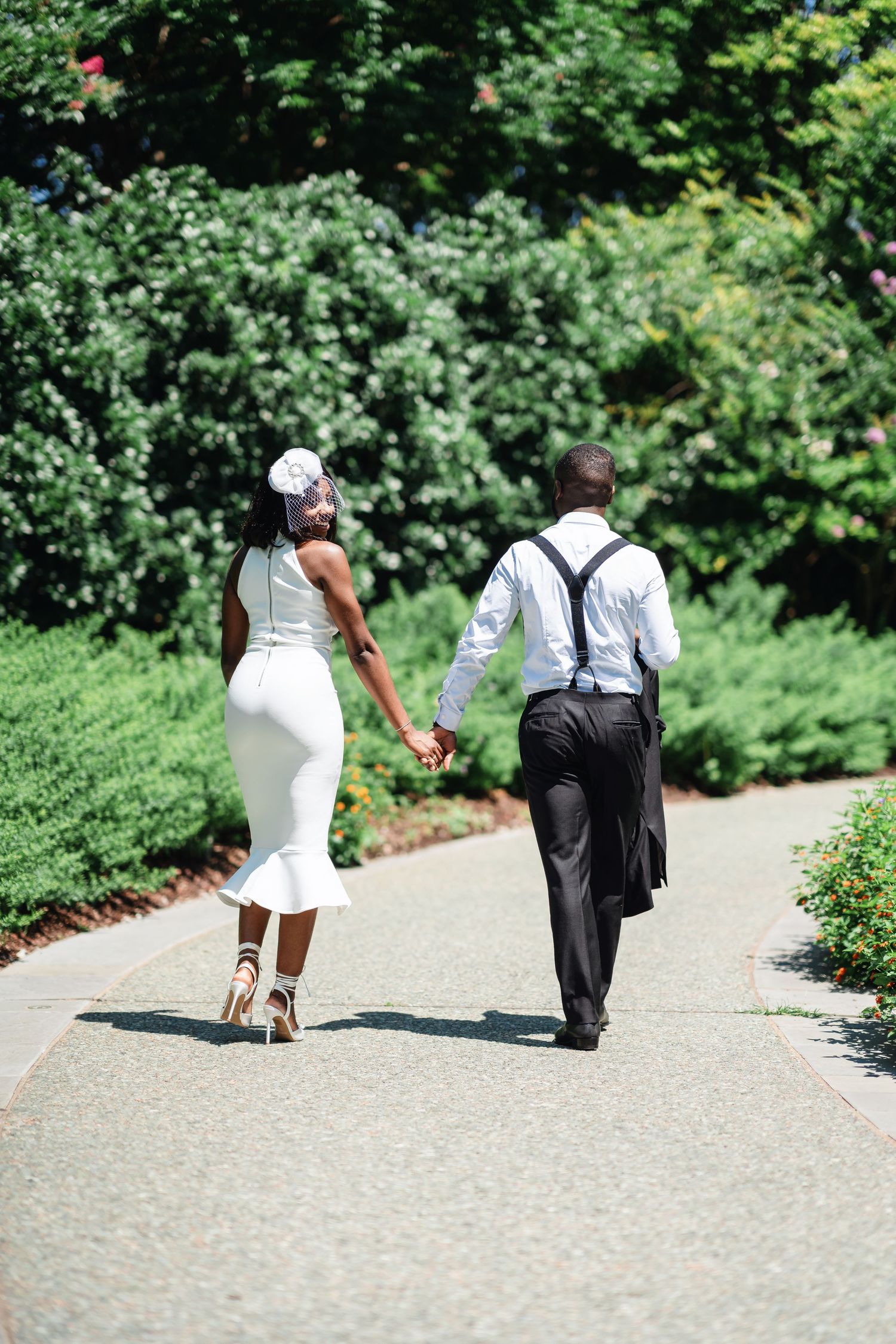 A couple walks hand in hand down a garden path surrounded by green landscaping.