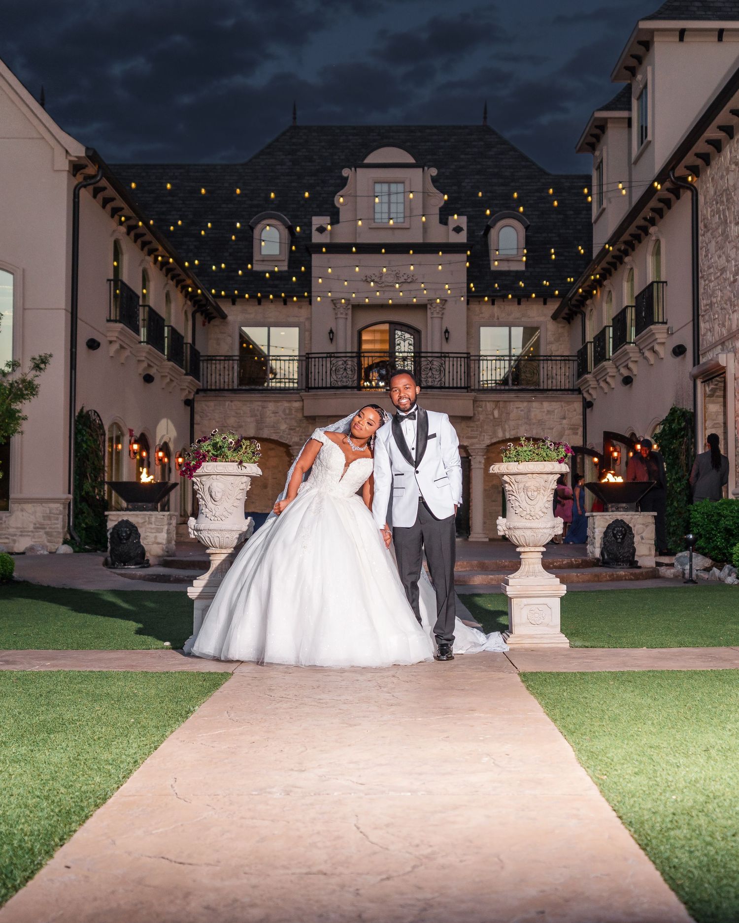 Wedding party poses for photos in an elegant outdoor courtyard venue with string lights and architectural details.