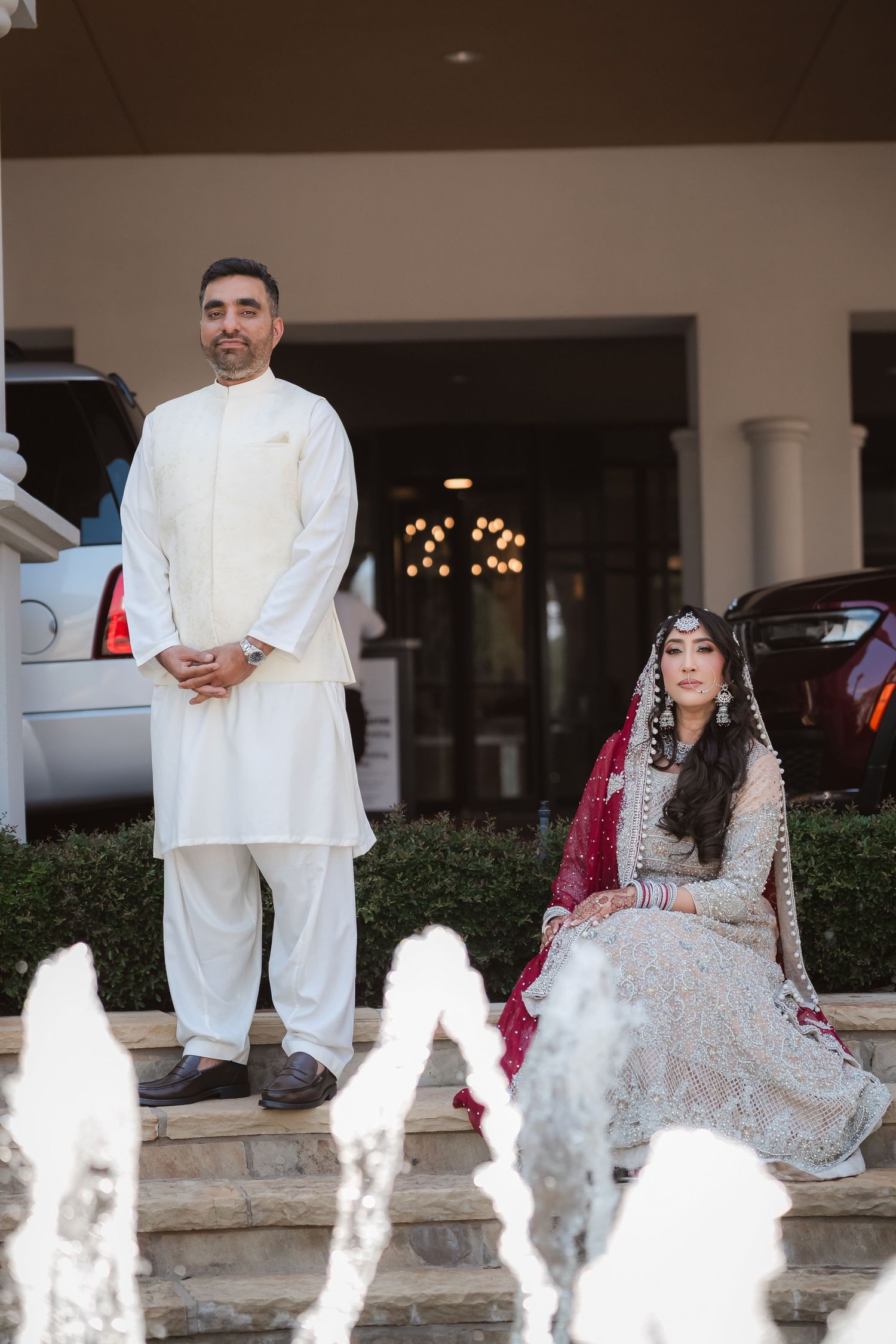 Wedding party seated by decorative fountain outside elegant building entrance.