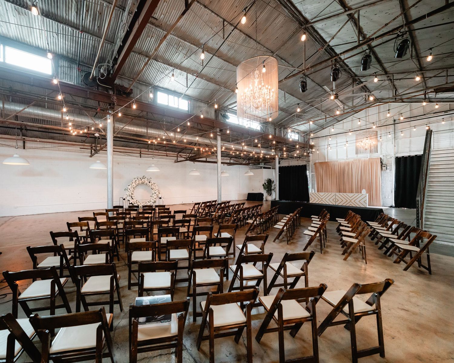 Industrial wedding ceremony space with wooden cross-back chairs arranged in rows under exposed beams.