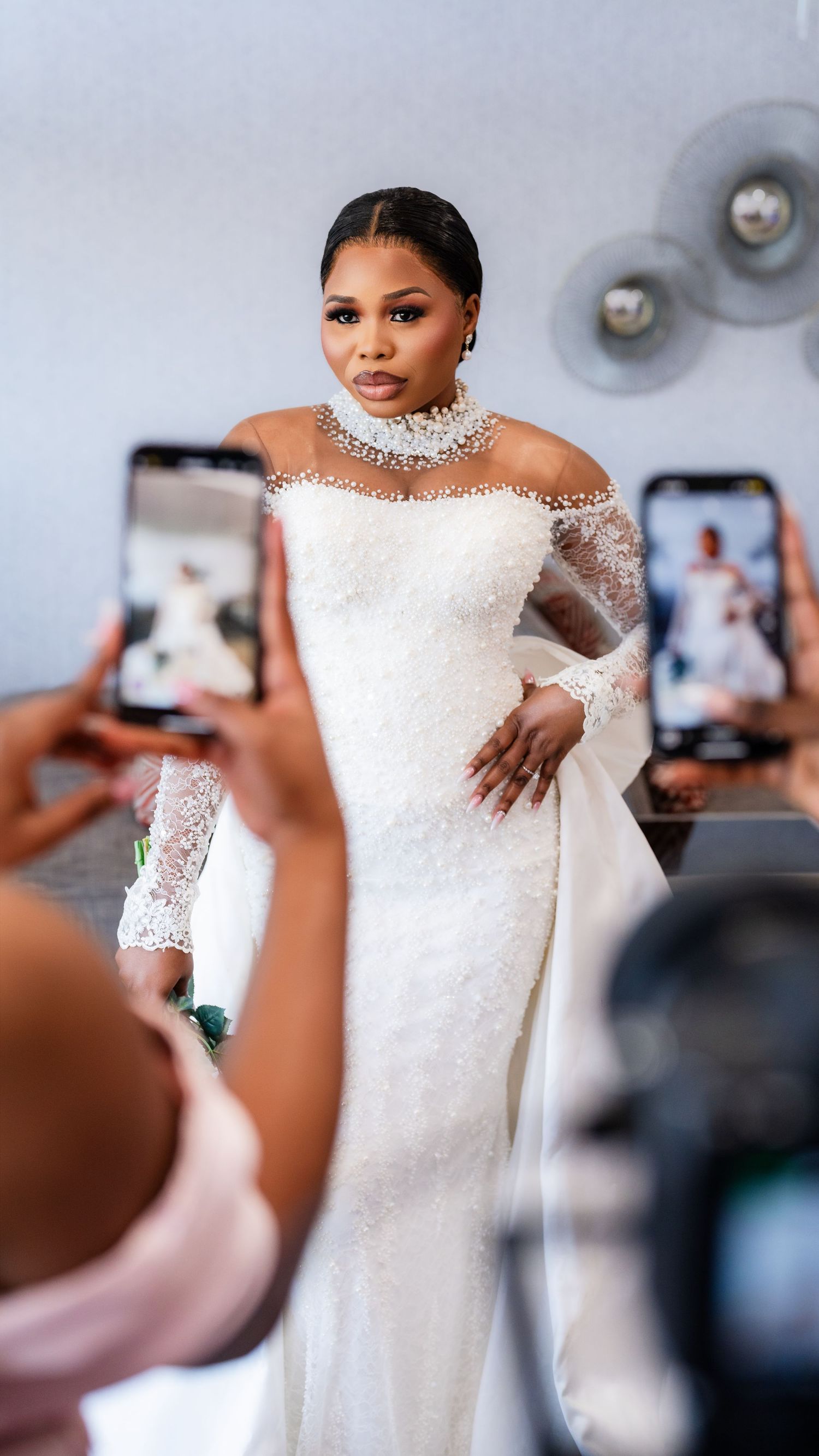 A bride poses in an elegant off-shoulder white wedding gown while guests take photos on their phones.