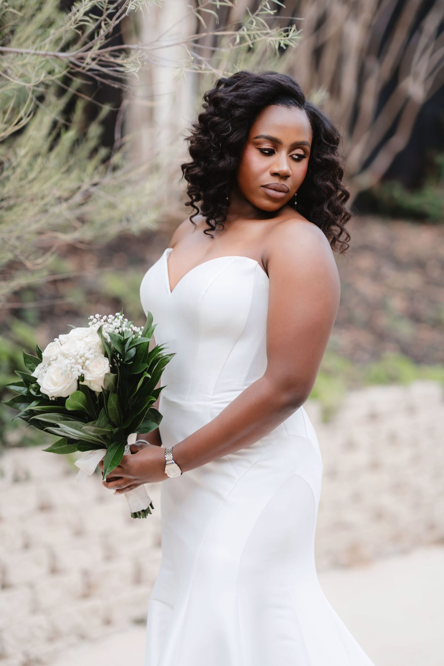 A bride in an elegant white wedding dress holds a bouquet against a natural outdoor backdrop.