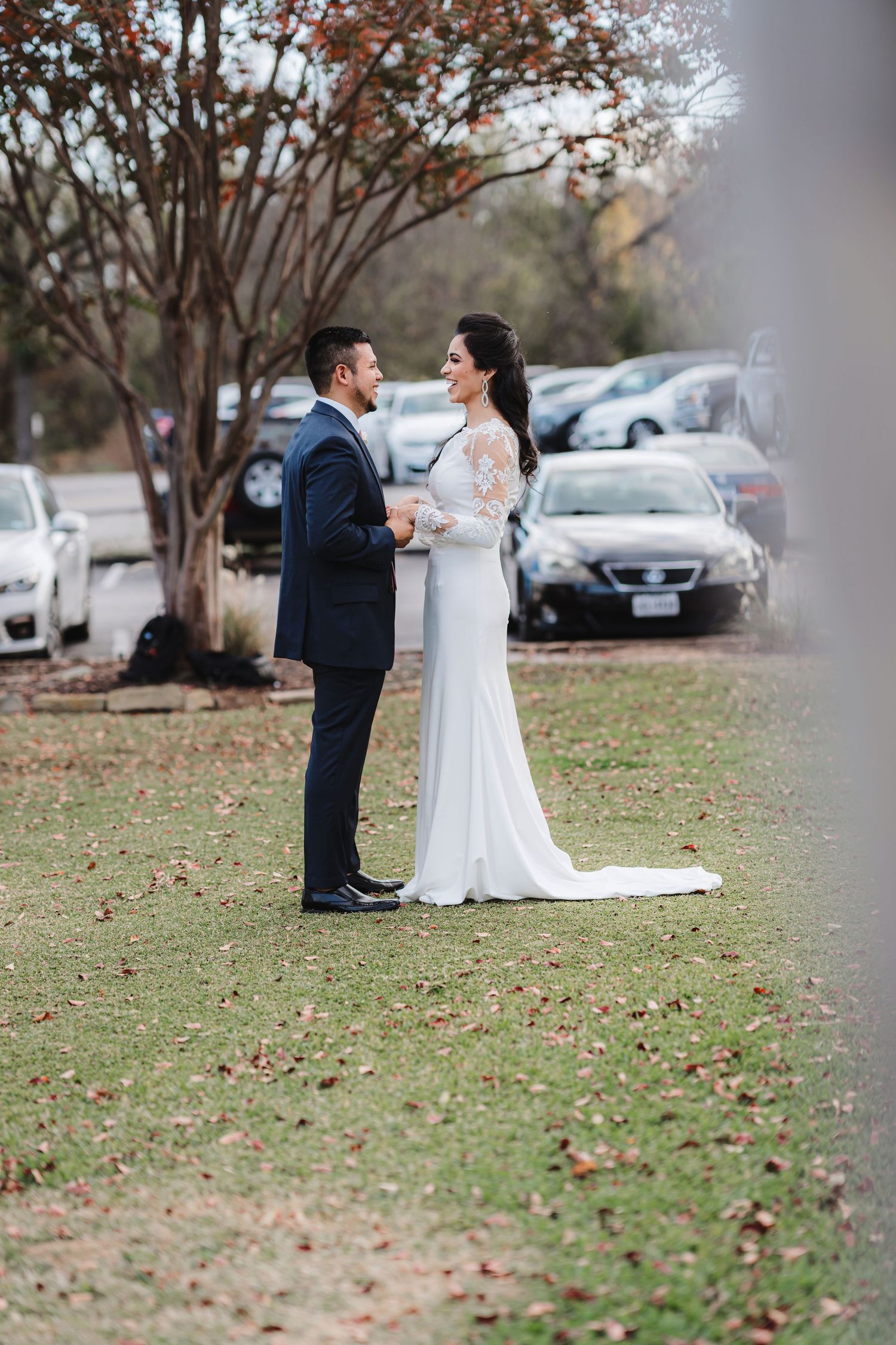 Newlyweds share a romantic moment on a lawn with autumn trees in the background.