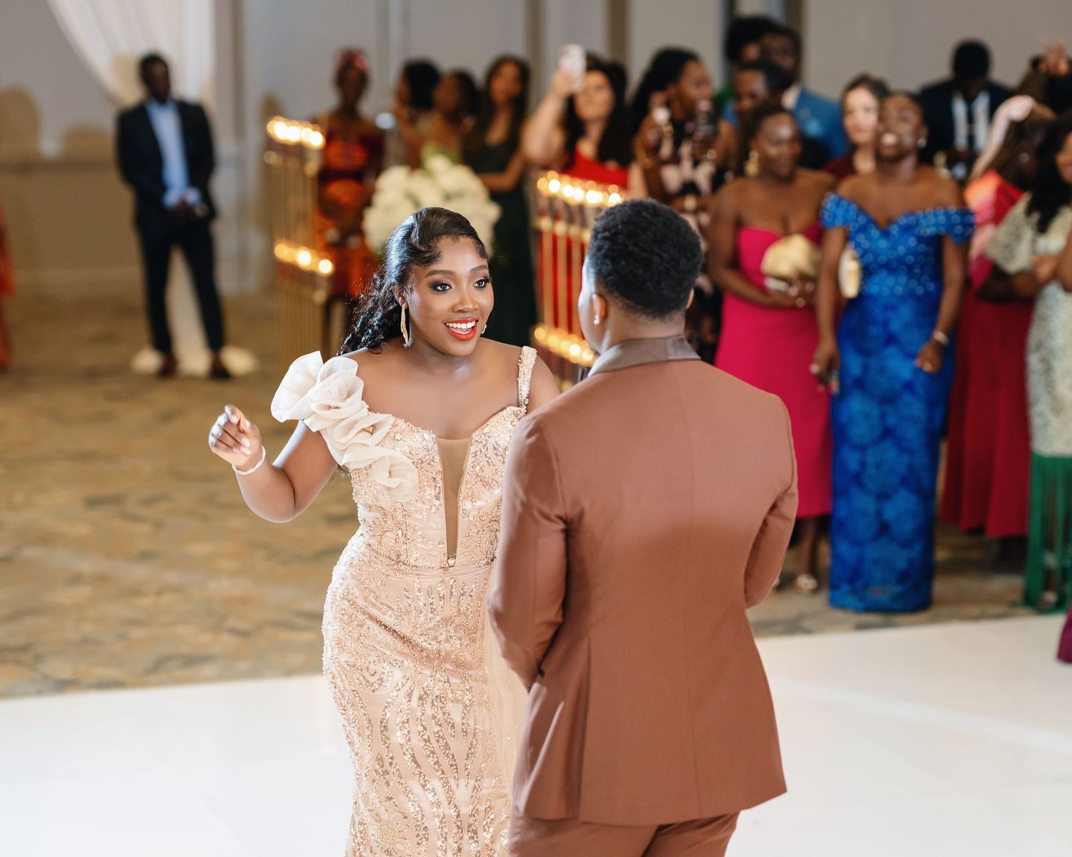 A couple dances at their wedding reception while guests in colorful attire watch from the background.