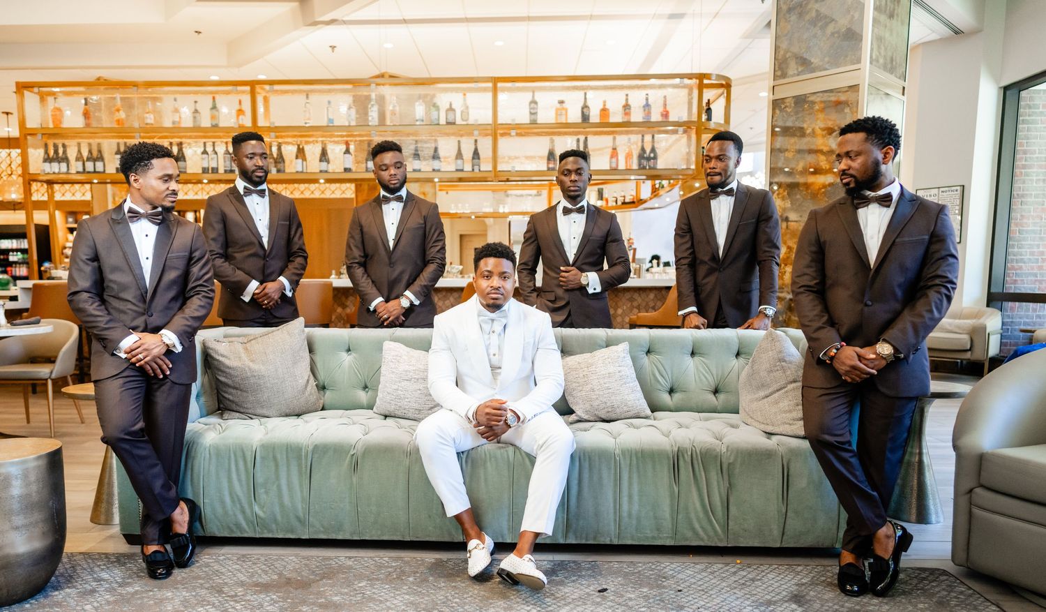 Groomsmen in black tuxedos and bow ties sit casually on gray couches in a modern lounge area.